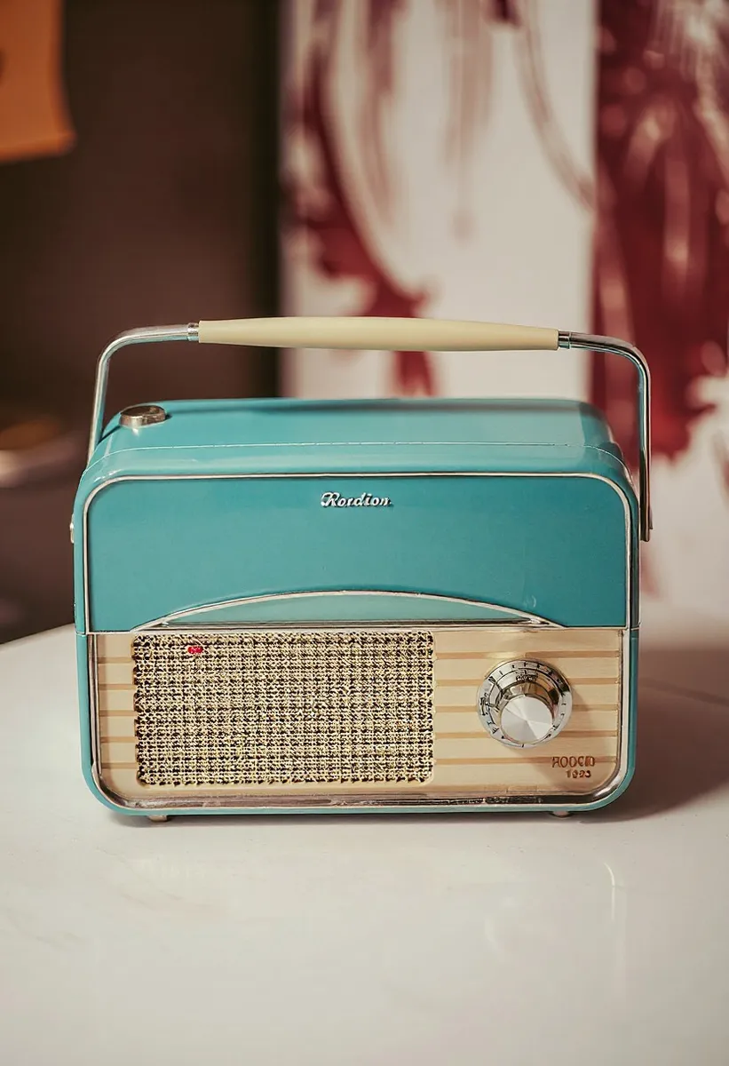 Turquoise retro radio with a beige woven speaker grille, analog dial knob, and cream-colored handle on top, placed on a white surface.