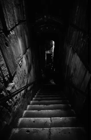 Black and white photograph of a narrow, worn stone staircase with metal handrails leading down into a dark, arched stone tunnel with a mysterious rectangular opening at the bottom.