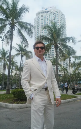 Tall man wearing a white pinstriped suit and sunglasses stands confidently in front of palm trees and a modern Miami skyscraper, shot from a low camera angle.