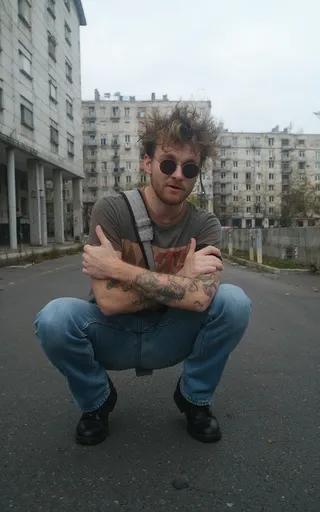 90s European DJ with spiky hair wearing black shades, hipster outfit, and jeans, crouched with arms crossed on a street with concrete buildings in the background