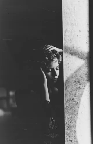 Black and white photograph of a woman with short wavy hair standing in a dimly lit room with eyes closed and hands on her head, showing a moody introspective atmosphere with vintage grainy texture and soft cinematic light from upper left.