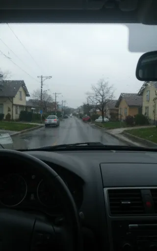 View through a car windshield during a rainy drive in a suburban neighborhood with American style houses and wet roads, taken with a slight motion blur.