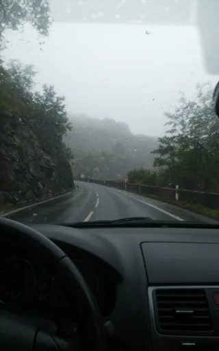 View from inside a car driving on a wet, foggy winding road through a cliffy, mountainous region in Scotland with rain droplets visible on the windshield.