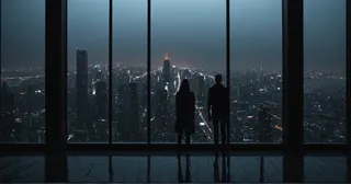 Two silhouetted figures stand inside a dark interior, framed by floor-to-ceiling windows, overlooking a foggy cityscape illuminated by neon lights and tall skyscrapers at night.