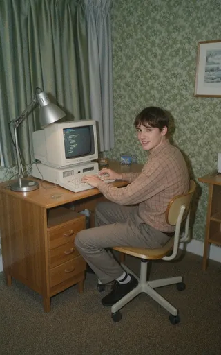 A college student typing code on a 1990s vintage computer in a green wallpapered dorm room with carpeted floors and a wooden desk, captured with nostalgic grainy texture.