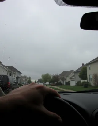 View from inside a car driving through a rainy suburban neighborhood, showing a hand on the steering wheel and raindrops on the windshield.