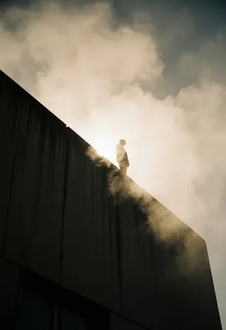 A silhouette of a person standing on the edge of a fog-covered brutalist rooftop with smoke swirling, backlit by a warm beam of sunlight piercing through the haze.