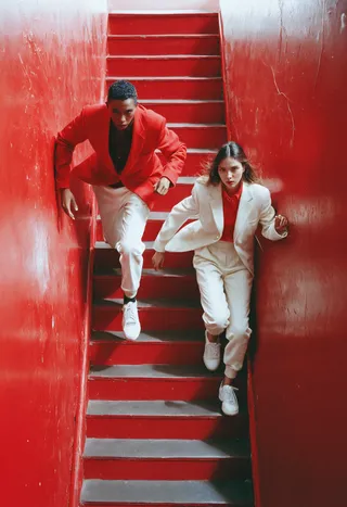 Overhead studio shot of a male and female model climbing a red staircase, wearing contrasting red sport coats and white sweatpants, in a Vogue-style fashion cover.