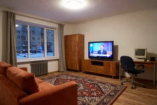 Retro Soviet style living room with an orange sofa, wooden cabinets, TV showing news, vintage computer on desk, Persian carpet, snowy view outside window at night.