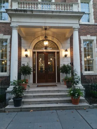 Traditional New York brownstone entrance featuring double arched wooden doors, warm hanging lantern lighting, white columns, and potted plants on stone steps.