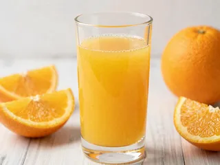 A clear glass filled with fresh orange juice surrounded by orange slices and a whole orange on a light wooden surface.