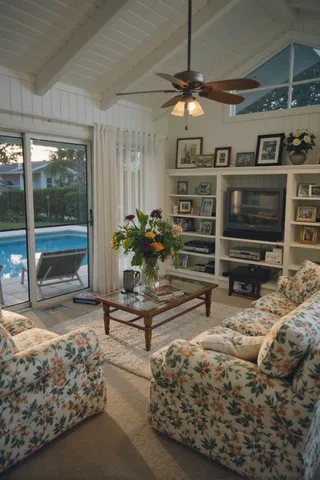 A 1980s Florida style living room featuring floral sofas, sliding glass doors leading to a pool in a screened area, built-in shelves with vintage electronics and photo frames, during golden hour.