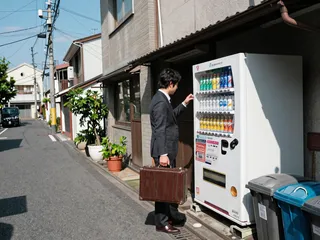 A man in a dark suit holding a brown briefcase buying a soda from a white Japanese vending machine along a quiet Tokyo backstreet with potted plants and trash bins.