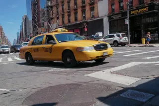 Close-up of a yellow 1990s New York City taxi cab driving on an urban street with sunny blue sky and surrounding buildings.