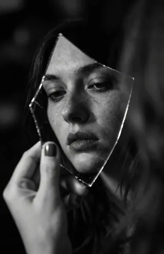 Close-up monochromatic portrait showing a feminine hand holding a jagged fragment of a shattered mirror reflecting a melancholic woman's face with dramatic chiaroscuro lighting.