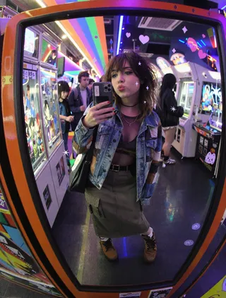 A young woman in a patchwork denim jacket takes a playful mirror selfie in a neon-lit arcade filled with retro game machines and colorful kawaii stickers on the mirror.