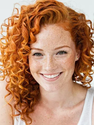 Close-up portrait of a smiling woman with curly orange hair and freckles against a white background