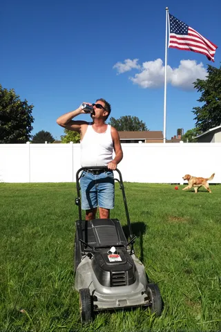 Man in jean shorts and white tank top drinking a can of beer while pushing a lawn mower in a suburban yard with a white fence, American flag waving, and a golden retriever chasing a ball.