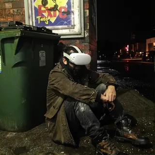 Dystopian cyberpunk scene with a homeless man wearing a white Oculus Quest VR headset sitting on a wet concrete road next to a green trash container and brick wall covered with graffiti at night after rain.