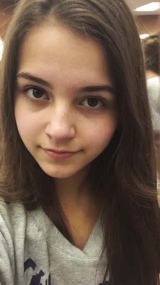 Close-up selfie of a college girl with long brown hair wearing a grey casual top, taken with an iPhone.