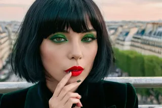 Close-up photograph of a French woman with black hair in a pixie cut, wearing bright red lipstick and green eyeshadow, smoking a cigarette with a blurred Paris cityscape background.