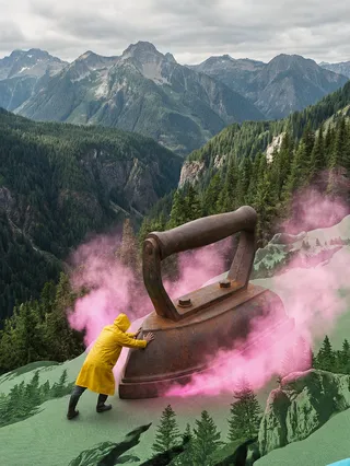 A mountain landscape where a person in a yellow raincoat pushes a colossal rusty iron pressing a green textile terrain, surrounded by pink steam and dense pine forests.