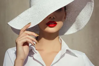 Close-up of a woman wearing a wide-brimmed white sun hat tilted diagonally, casting a shadow over part of her face. She has vibrant red lipstick, smooth matte skin, and metallic gold almond-shaped nails, dressed in a crisp white collared shirt.