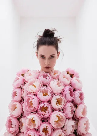 Model with messy single hair bun wearing a dress completely covered with pink peonies in a white minimalist room, shown from the front view.