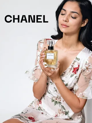 Young woman with black hair wearing a white floral dress holding a perfume bottle with closed eyes and sitting against a white background.