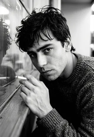 Moody black and white portrait of a young man with tousled dark hair, leaning on a reflective surface, holding a lit cigarette, wearing a textured knitted sweater, with a melancholic gaze.