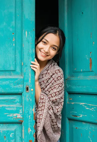 A young woman smiling warmly as she playfully peeks from behind a vibrant, weathered turquoise wooden door, wrapped in a brown and white geometric patterned fabric.