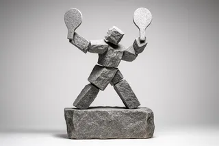 Minimalist Nordic-style sculpture of a tennis player made from rough gray granite blocks, posed mid-swing with two stone rackets, standing on a granite base.