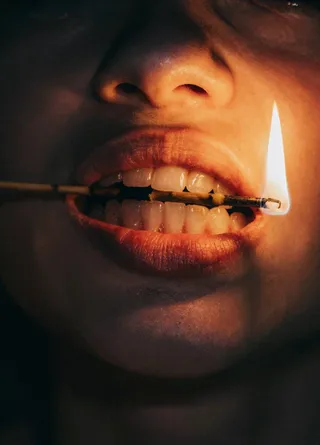 Close-up of a woman's mouth holding a burning flame horizontally between her teeth, illuminated by warm orange-yellow light highlighting red lips and smooth skin texture against a dark background.