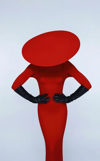 A confident woman stands with hands on hips, wearing a saturated scarlet dress and a wide, dramatic red hat, complemented by long black leather gloves against a flat white background.