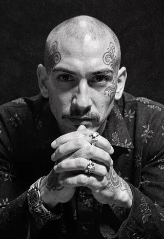 Close-up black and white portrait of a shaved-head man with intricate head tattoos and a faint mustache, clasping his tattooed hands adorned with rings and a patterned bracelet, under dramatic, film grain lighting.