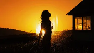 Silhouette of a woman partially obscured by vertical light streaks standing in a field near a cabin at sunset with warm golden and orange hues.