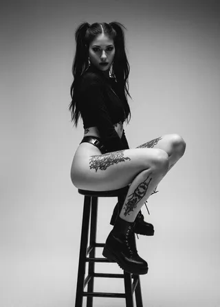 Black and white photograph of a punk woman with long wavy hair in pigtails, sitting on a tall black wooden stool. She has multiple tattoos on her thighs, wears a black cropped top and knee-high lace-up combat boots. The image has grainy texture and moody lighting.