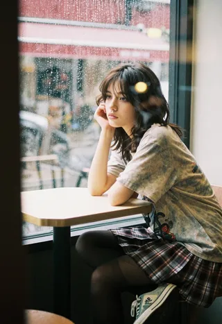 Young woman with dark wavy hair sits pensively at a cafe table by a rain-speckled window, wearing a muted plaid skirt and vintage tee in soft 90s grunge indie style.