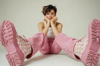Young woman with short pixie haircut wearing a white tank top and wide-leg pink corduroy pants, sitting on a white floor in a minimalist studio. She rests her chin on her hands with elbows on the floor, with large chunky pink boots prominently in the foreground.