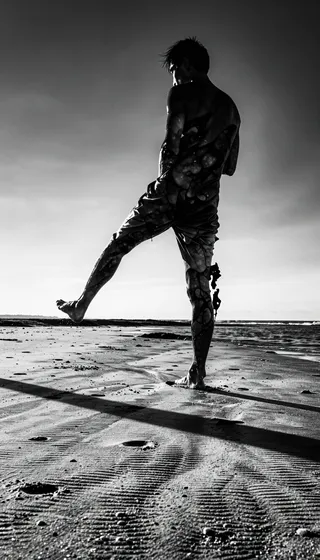 Black and white high-contrast photograph of a dynamic figure standing on wet sand at the beach, lifting one leg, with a long sharp shadow cast on the rippled sand surface.