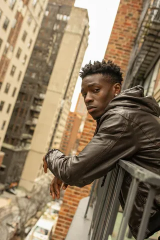 Young African American man with a serious expression in a leather hooded jacket leaning on a metal balcony railing with a tilted cityscape background.