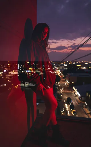 A young woman in an oversized hoodie and boots stands against a red-lit wall, with intense neon lighting and a double exposure effect merging city neon signs and street lights in the background.