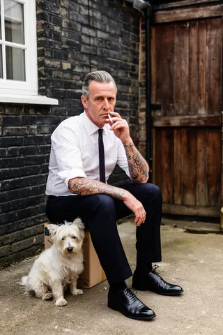Middle-aged Englishman with graying swept-back hair and heavily tattooed arms, wearing a white shirt and dark trousers, sitting on a box in an outdoor backyard smoking a cigarette, with a small white terrier dog beside him, against an old brick wall and wooden door.