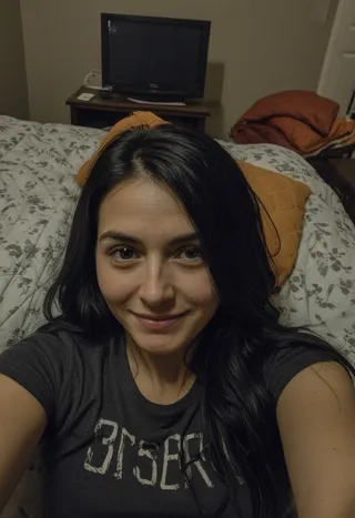 A woman with long black hair wearing a t-shirt takes a selfie in a dimly lit bedroom at night, lying on a bed covered with comforters and quilts.