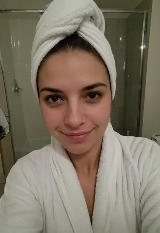 A woman wearing a white towel turban and white bathrobe, smiling slightly while taking a selfie in a bathroom.