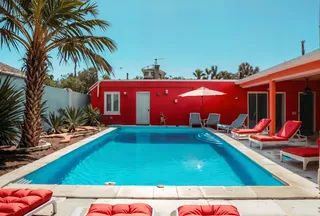 A symmetrical swimming pool scene with crystal clear blue water, surrounded by red cushioned loungers, palm trees, and a red building under a clear blue sky.