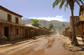 A scenic view of an 1800s colonial Spanish street featuring a muddy road, wooden huts, palm trees, horses, and rustic shops under a sunny sky.
