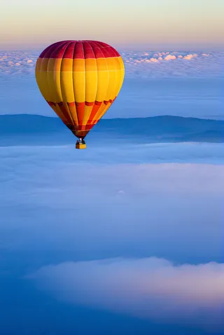 Colorful hot air balloon floating above clouds during early morning sunrise with serene blue skies and distant mountains.