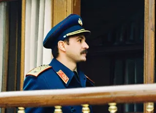 Side profile of a Soviet police officer wearing a blue uniform and cap with red and gold insignia, standing near a wooden railing in front of a window