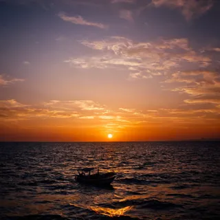 A cinematic sunset over the ocean showing a small boat silhouette on water with vibrant orange sky and dramatic clouds.